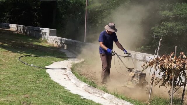 Wide shot of a male worker steering a heavy motor tiller on dry dirt near a stone wall, creating dust while preparing soil for planting.