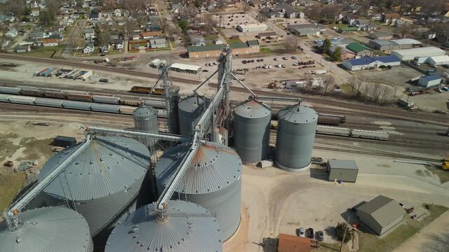Ascending drone view reveals a large grain storage complex with metal silos, conveyor systems and freight trains on parallel railway tracks
