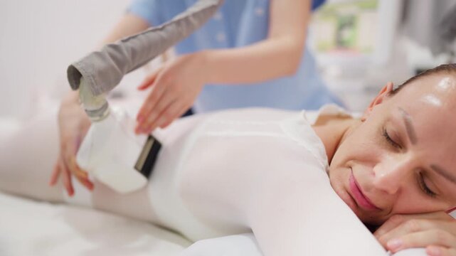 Young woman receiving an lpg massage procedure on her back and buttocks in a beauty salon, with a therapist using a modern vacuum roller machine for body shaping and skin toning