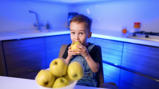 Boy biting into apple in blue lit kitchen. A child sits at a counter beside a bowl of apples under vivid blue ambient lighting.