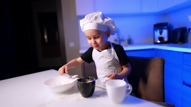 Boy in toque scooping white flour with spoon into cup on kitchen table. Ambient blue illumination shines on cabinetry while a child cooks indoors.