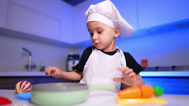 Young child wearing chef hat standing behind toy cookware on white counter. Bright artificial blue lighting illuminates a modern home kitchen during playtime.