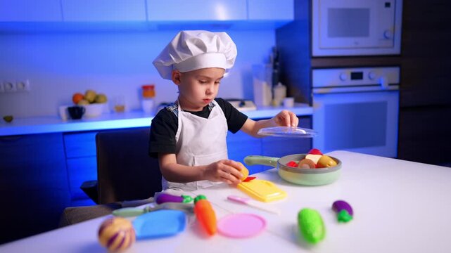 Boy in chef costume playing with colorful toy vegetables and plastic pan. Fake carrots and corn rest on a white surface lit by ambient blue background light.