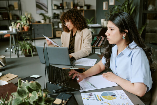 Diverse young adult female colleagues working on laptops in modern office, analyzing financial charts. Image suitable for corporate communication and remote hybrid work marketing