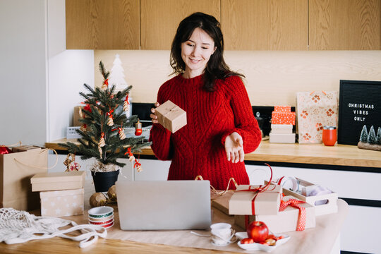 Woman holds kraft wrapped parcel while working on laptop among gifts and wrapping supplies. Christmas small business laptop, managing orders christmas season, small business holiday computer work
