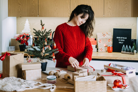 Woman ties ribbon on kraft box at cluttered kitchen table near CHRISTMAS VIBES ONLY sign. Christmas rush preparation, holiday overwhelm concept, juggling christmas tasks
