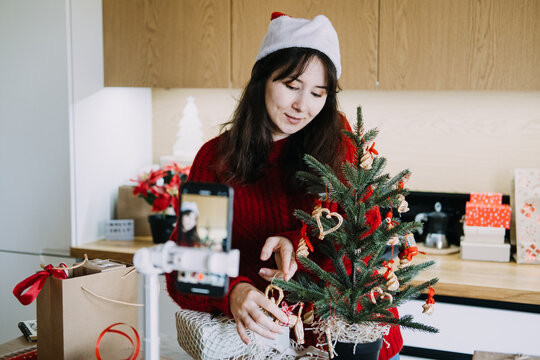 Woman in Santa hat decorates small potted tree while phone on tripod records her in kitchen. Filming christmas vlog phone, content creator holiday video, recording festive video content