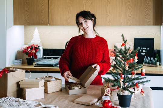 Woman holds wooden brush over open kraft box in kitchen near CHRISTMAS VIBES ONLY sign. Preparing christmas order for shipment, assembling product for customer, small shop order preparation process