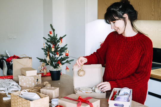Smiling woman places paper ornament on open kraft box with brush and jar in kitchen. Sustainable christmas gift set, plastic free gift packaging, handmade spa box preparation