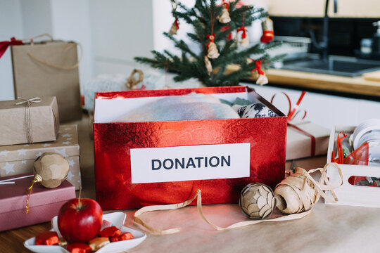 DONATION box filled with knitwear and heart stands on table among wrapped gifts near Christmas tree. Winter clothes donation drive, holiday charity collection box, donate warm clothing christmas