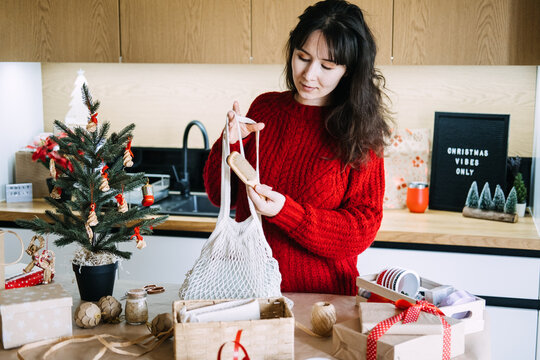 Woman holds mesh bag and wooden brush over gift basket in kitchen near CHRISTMAS VIBES ONLY sign. Content creator christmas gift, filming christmas gift assembly, small business christmas gift content