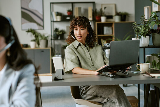Biracial young adult woman working in call center wearing headset and using laptop. Image suitable for customer support, telemarketing, and corporate communication marketing materials