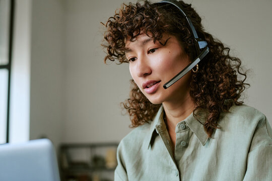 Biracial young adult woman wearing headset working in call center, talking to client on laptop. Image represents customer support, telemarketing, and remote helpdesk services, close up