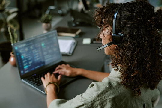 Young adult woman working in call center wearing headset and typing on laptop. Over shoulder view of female operator providing customer support for telecommunications marketing