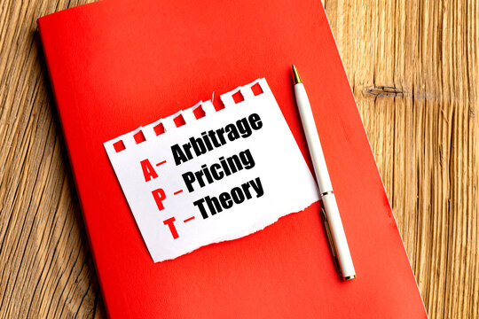 Arbitrage pricing theory concept with apt acronym on paper, red notebook and pen on wooden desk, representing finance