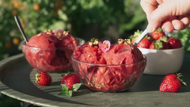 A beautiful close-up shot of a hand using a small spoon to take a scoop of bright red homemade strawberry sorbet. The cold dessert is served in elegant glass bowls on a metal tray