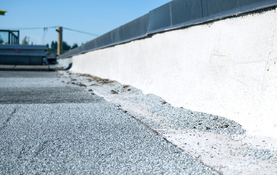Flat roof parapet with granule loss on modified bitumen cap sheet. Low angle view. Aggregate pile visible at base. SBS 2-ply roofing system requiring maintenance. Selective focus.