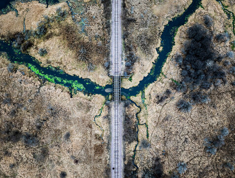 Overhead top-down aerial drone view of a straight railroad track and bridge crossing a winding dark blue river with patches of green algae. Abstract infrastructure and nature landscape.