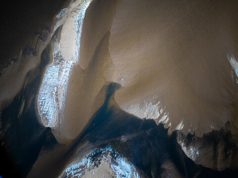 Overhead top-down aerial drone view of smooth brown sandbars meeting dark water channels with pockets of white ice textures. Moody abstract natural river landscape background photography.