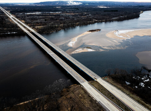 High-angle aerial view of a dual-span concrete highway bridge crossing a wide river with winter sandbars and ice patches. Overhead transportation infrastructure drone photography.