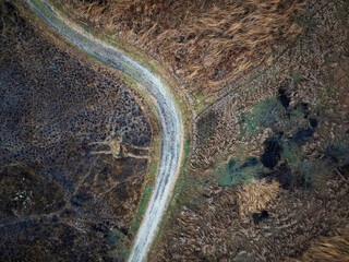 Overhead aerial drone view of a curved dirt road sweeping through a dark, textured rural landscape with patches of dry brown grass and brush. Top-down scenic wilderness photography. © Jason