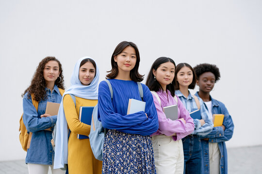 Group of diverse female students standing together, wearing casual clothing and holding notebooks, with a plain white wall in the background