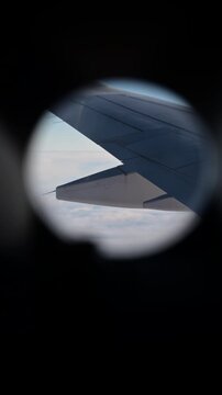 Vertical view through a round cabin window of an airplane wing above a white cloud layer