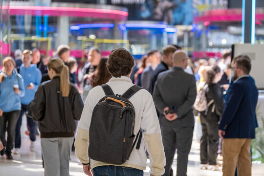 Backpack-wearing visitor walks into a crowded exhibition hall filled with people and colorful illuminated booths.