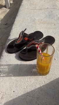 Vertical HD close-up video of handcrafted African beaded leather sandals (flip-flops) lying on a sunny wall on Paje beach, Zanzibar. Next to them stands a glass of fresh tropical orange/mango juice.