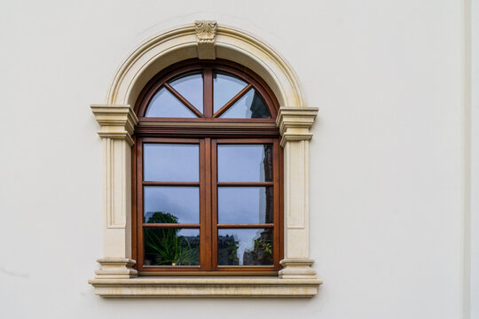 A classic architectural window with a rich brown wooden frame, a rectangular grid section paired with an adjacent arched fanlight, all set within an ornate, stepped beige masonry, stucco surround. 