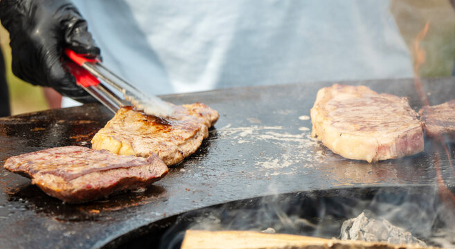 Black gloves handle tongs over sizzling meat