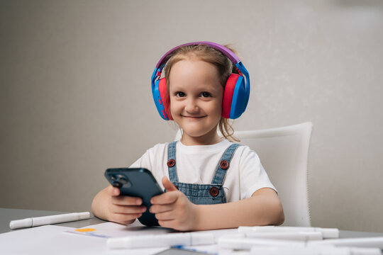 Studio portrait of young girl smiling while wearing colorful headphones and holding smartphone, concept of childhood, leisure, modern technology, online content consumption in bright setting.