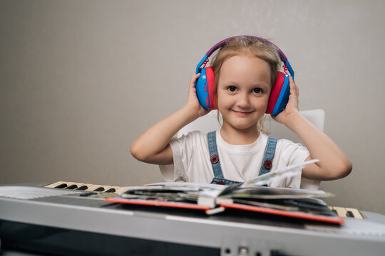Little girl smiling at camera, wearing colorful headphones and sitting in front of digital piano or synthesizer with music book open, enjoying music lesson.