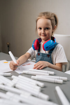 Young girl coloring on paper with marker, wearing an overall dress and blue and red headphones around neck, focusing on creative development and childhood learning at home.