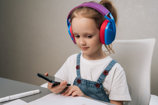 Young child wearing colorful headphones and denim overalls looking intently at screen of mobile phone while sitting at desk, engaging with digital media.