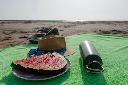 Fresh sliced watermelon with hat and metal water bottle on beach