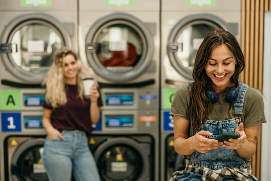 Happy women friends using smartphone at self-service laundromat