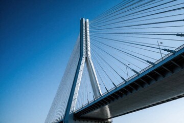 Fototapeta premium Modern cable stayed bridge structure with massive white pylon, steel cables against clear blue sky. Geometric lines of suspension wires create symmetry. Low angle view of engineering marvel overpass