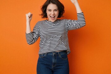 Fototapeta premium Woman full-length. Exhilaration, arms raised, hands together, sporting denim, long-sleeve striped shirt. Festivity, celebration, business triumph, gladdened look, happy energy. Brilliant setting,