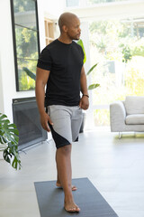 Fototapeta premium African American man standing barefoot on dark exercise mat in bright living room, wearing bracelet