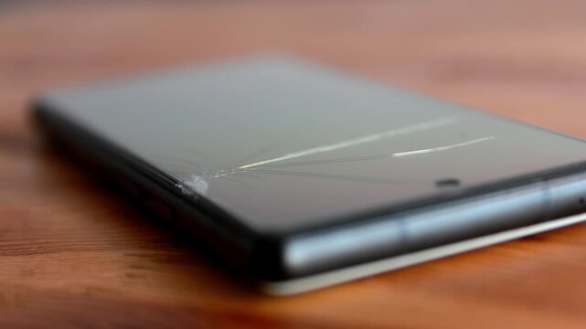 Close-up of a modern smartphone with a severely cracked screen on a wooden table. Close-up of a modern smartphone lying on a wooden table with a cracked display glass and visible screen damage