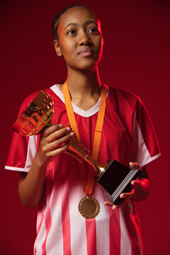 Female athlete wearing red white jersey and studs, holding gold trophy with medal ribbon in studio
