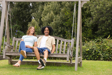 Naklejka premium Couple sitting on wooden swing in backyard, showing metal chains, casual shirts and shorts