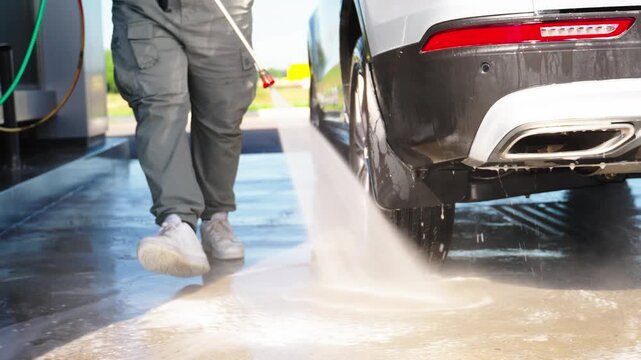 Water blasts clean a car's rear end. Worker stands near the vehicle. The ground is wet with foam and spray. Bright sun illuminates the scene. Ideal for automotive or cleaning business ads