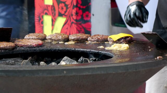 Grilled burgers sizzle over glowing charcoal. Chef's gloved hand places cheese slices. Fatty patties cook in open flame. Food vendor prepares quick meal. Perfect for food festival or street market ads