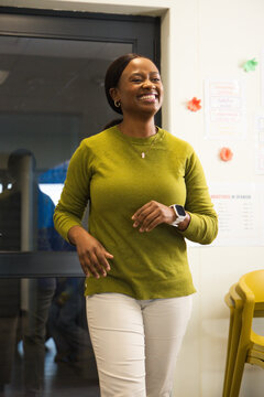 Adult African woman walking in classroom, speaking near glass door with posters and yellow chairs