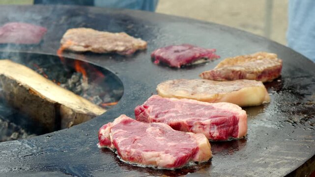 Steaks sizzle on a circular grill. Fire flickers beneath the cooking meat. Grill surface shows signs of heavy use. Heat rises from the sizzling meat. No people are visible in the frame