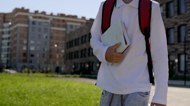 Student walks toward campus, holding books. Backpack straps show casual, focused posture. Bright sunlight highlights brick buildings and green grass