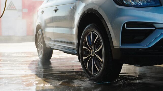 Water sprays clean sleek silver SUV. Wet pavement reflects car's glossy finish. Rinsing wheels suggests recent cleaning. Vehicle gleams under soft, diffused light