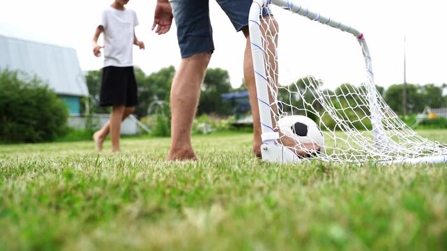 Footage shows two barefoot figures near a small goal. One adult stands close to the net, holding a ball. Child runs behind, eager to play. The grass is green and slightly uneven